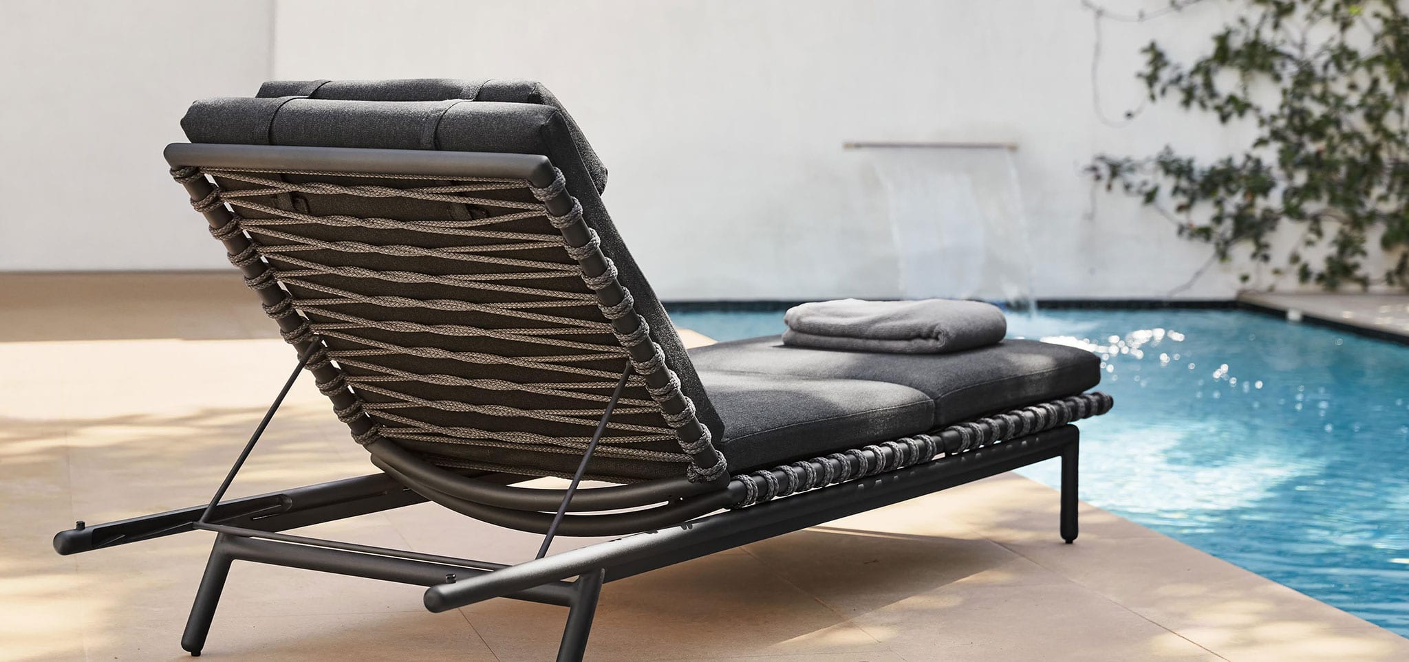 Oscar chaise with dark colored thick cushions from Brown Jordan has a towel folded on it as it sits on a patio facing a luxurious pool
