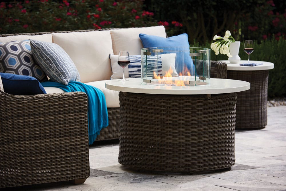 Oasis fire pit set and patio furniture set with half-full wine glasses on the drink tables from Lane Venture on a stone patio with a woodlands in the background